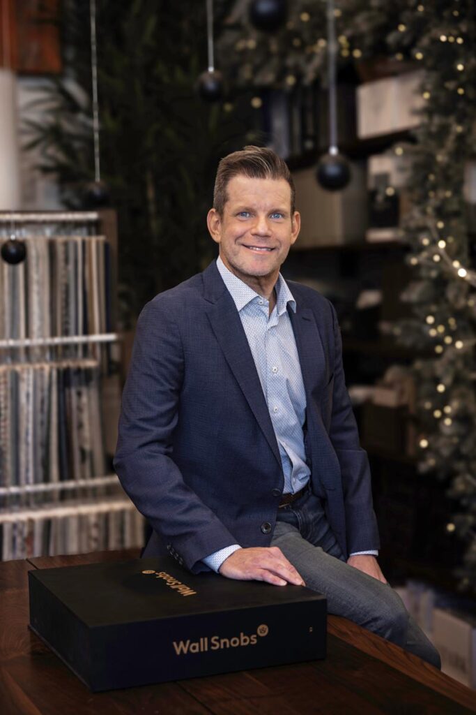 Smiling man in a suit sitting on a table, surrounded by fabric samples and decorations © Nashville Interiors